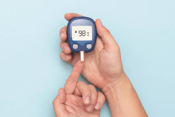 Person using digital blood glucose meter to test blood sugar level via fingertip on blue background
